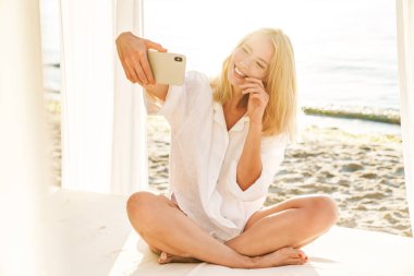 Yazın kumsalda oteldeyken cep telefonuyla selfie çeken neşeli sarışın bir kadın.