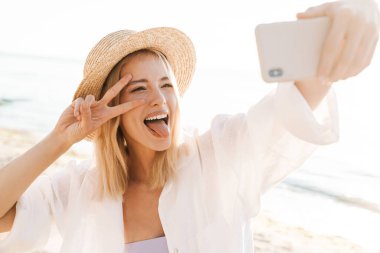 Mutlu bir kadının yaz plajında dururken cep telefonuyla selfie çektiği görüntüsü.