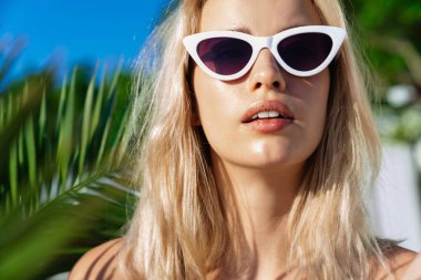 Güneş gözlüklü çekici sarışın bir kadının yaz kumsalında kameraya baktığı görüntü.