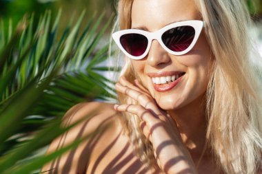 Güneş gözlüklü çekici sarışın bir kadının yaz plajında gülümseyen ve yan bakan görüntüsü.