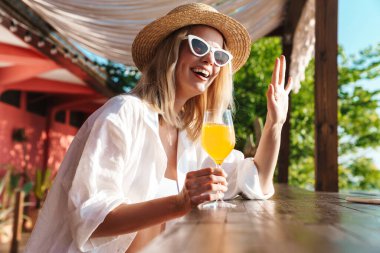 Plaj barında kokteyl içerken gülümseyen ve el sallayan sevimli bir kadın resmi.