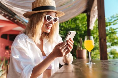 Plaj barında kokteyl içerken gülümseyen ve cep telefonu kullanan çekici bir kadın görüntüsü