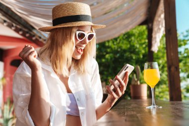 Çekici, neşeli bir kadının sahil barında kokteyl içerken cep telefonu kullanması.