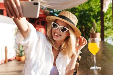Plaj barında kokteyl içerken cep telefonuyla selfie çekerken gülümseyen neşeli bir kadın görüntüsü.