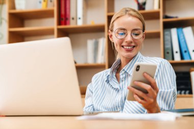 Ofiste otururken dizüstü bilgisayar ve cep telefonuyla çalışan mutlu genç bir kadın görüntüsü