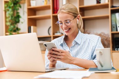 Ofiste dizüstü bilgisayarla çalışırken kulaklık ve cep telefonu kullanan gülümseyen genç iş kadını görüntüsü