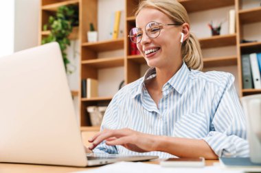 Ofiste dizüstü bilgisayarla çalışırken kulaklık takan ve gülen mutlu bir kadın görüntüsü.