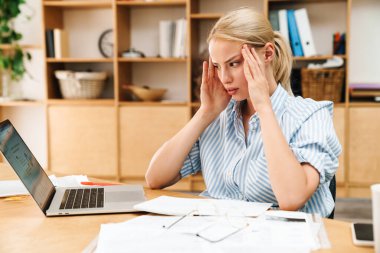 Ofiste çalışırken dizüstü bilgisayar kullanan, başı ağrıyan mutsuz sarışın bir kadın resmi.
