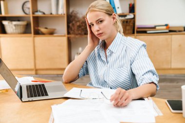 Ofiste otururken dizüstü bilgisayar kullanan, başı ağrıyan genç bir kadın resmi