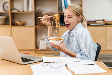 Makarna yiyen ve masada otururken dizüstü bilgisayarla çalışan heyecanlı genç bir kadın resmi.