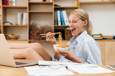 Makarna yiyen ve masada otururken dizüstü bilgisayarla çalışan heyecanlı genç bir kadın resmi.