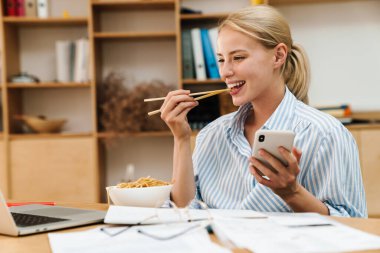 Ofiste dizüstü bilgisayar ve cep telefonuyla çalışırken erişte yiyen neşeli genç bir kadın görüntüsü