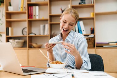Asya eriştesi yerken fotoğraf çeken ve bilgisayarla ofiste çalışan neşeli sarışın bir kadın resmi.