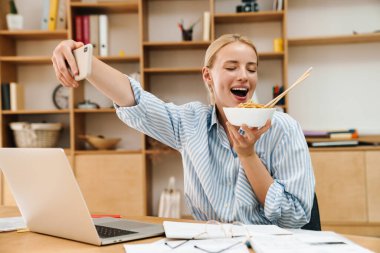 Asyalı erişte yerken, ofiste dizüstü bilgisayarla çalışırken selfie çeken neşeli sarışın bir kadın resmi.