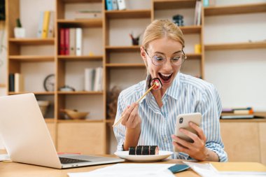 Ofiste cep telefonu ve dizüstü bilgisayar kullanırken yemek çubuklarıyla suşi yiyen sarışın bir kadın resmi