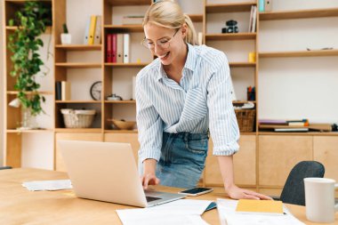 Ofiste otururken dizüstü bilgisayarla gülümseyen ve çalışan mutlu bir kadın resmi.