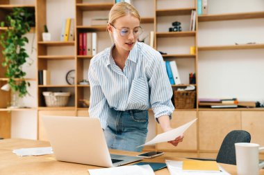 Ciddi, güzel bir kadının belge okuduğu ve masada otururken dizüstü bilgisayarla çalıştığı bir resim.