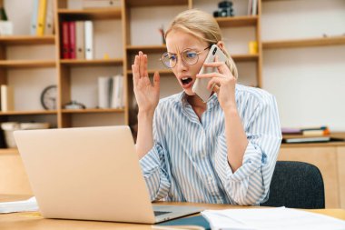 Ofiste dizüstü bilgisayarla çalışırken cep telefonuyla konuşan hoşnutsuz iş kadını görüntüsü