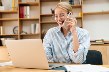 Ofiste dizüstü bilgisayarla çalışırken cep telefonuyla konuşan neşeli çekici bir iş kadını görüntüsü