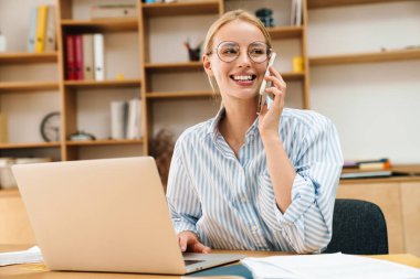 Ofiste dizüstü bilgisayarla çalışırken cep telefonuyla konuşan neşeli çekici bir iş kadını görüntüsü