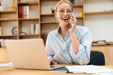 Ofiste dizüstü bilgisayarla çalışırken cep telefonuyla konuşan neşeli çekici bir iş kadını görüntüsü