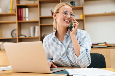 Ofiste dizüstü bilgisayarla çalışırken cep telefonuyla konuşan neşeli çekici bir iş kadını görüntüsü