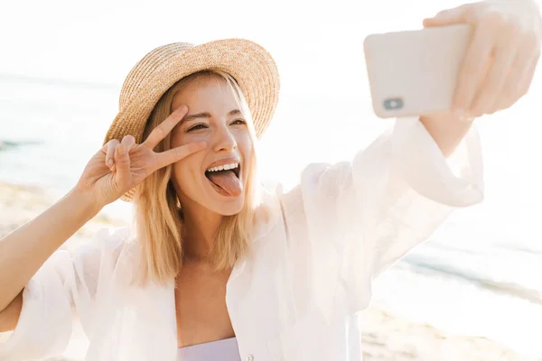 夏のビーチに立ちながら携帯電話で写真を撮り平和サインを示す幸せな女性のイメージ — ストック写真