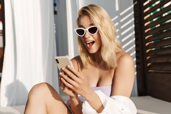 Image of seductive delighted woman in sunglasses using cellphone at hotel on summer beach