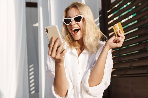 Image of excited woman in sunglasses holding credit card and using cellphone at hotel on summer beach