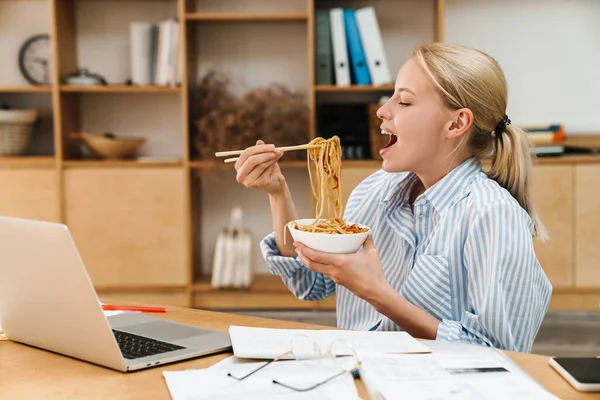 照片中快乐的金发女人一边工作一边拿着纸和笔记本电脑 一边吃着亚洲面 — 图库照片