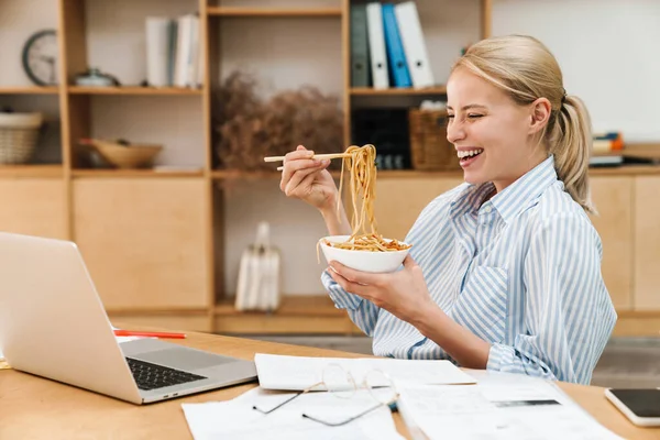 stock image Image of cheerful blonde woman eating asian noodle while working with papers and laptop in office