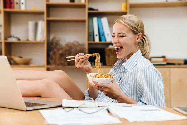Makarna yiyen ve masada otururken dizüstü bilgisayarla çalışan heyecanlı genç bir kadın resmi.