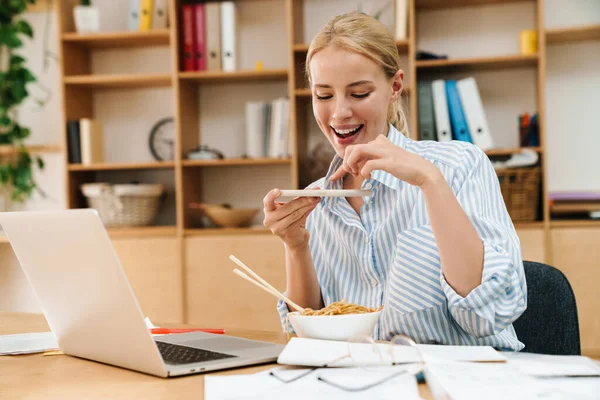 Asya eriştesi yerken fotoğraf çeken ve bilgisayarla ofiste çalışan neşeli sarışın bir kadın resmi.