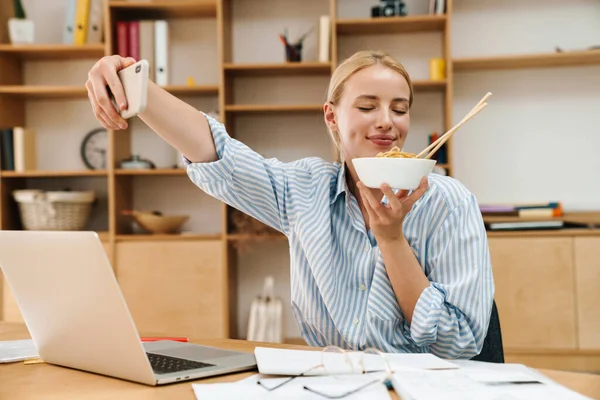 Asyalı erişte yerken, ofiste dizüstü bilgisayarla çalışırken selfie çeken neşeli sarışın bir kadın resmi.
