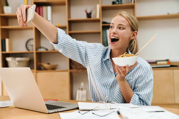 照片中快乐的金发女人一边吃亚洲面一边自拍 一边在办公室用笔记本电脑 — 图库照片