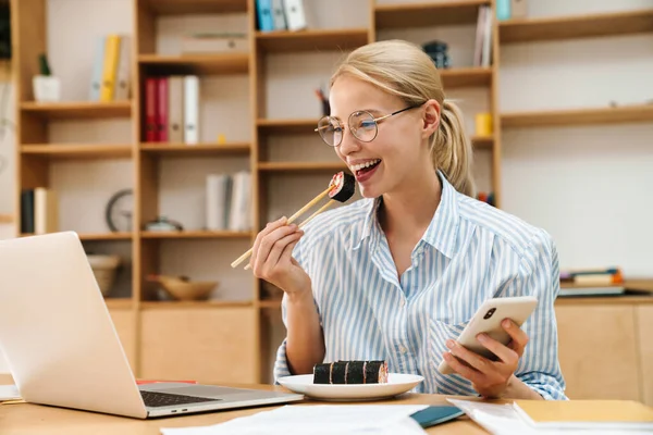 Ofiste cep telefonu ve laptop kullanırken yemek çubuklarıyla suşi yiyen mutlu sarışın kadın resmi