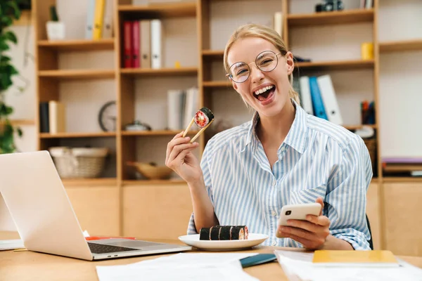 Ofiste cep telefonu ve laptop kullanırken yemek çubuklarıyla suşi yiyen heyecanlı sarışın kadın görüntüsü