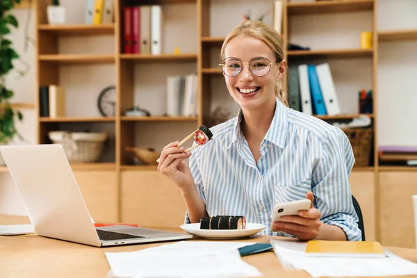 Ofiste cep telefonu ve laptop kullanırken yemek çubuklarıyla suşi yiyen mutlu sarışın kadın resmi