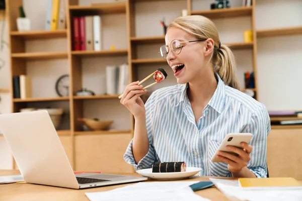 Ofiste cep telefonu ve laptop kullanırken yemek çubuklarıyla suşi yiyen mutlu sarışın kadın resmi