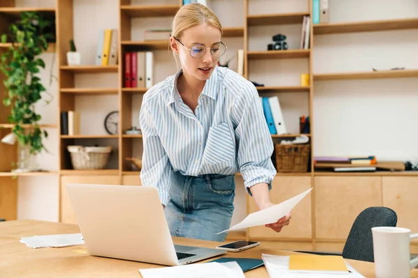 Ciddi, güzel bir kadının belge okuduğu ve masada otururken dizüstü bilgisayarla çalıştığı bir resim.