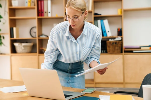 Ciddi, güzel bir kadının belge okuduğu ve masada otururken dizüstü bilgisayarla çalıştığı bir resim.