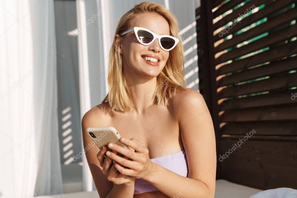 Image of seductive happy woman in sunglasses smiling and using cellphone at hotel on summer beach