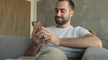 Genç pozitif adam cep telefonu kullanırken evde oturuyor.