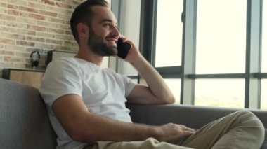 Genç, yakışıklı, neşeli adam cep telefonuyla konuşurken evde oturuyor.