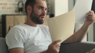Genç stresli bir adam, elinde belgelerle evde oturuyor.