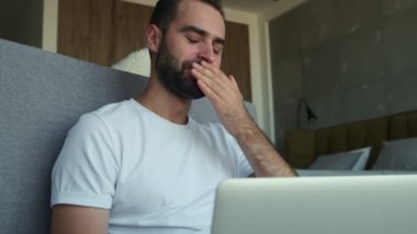 Genç esneyen adam evde oturuyor ve dizüstü bilgisayar kullanıyor.