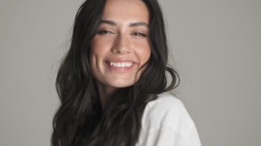 Gülümseyen genç bir kadın kameraya hava öpücüğü gönderiyor. Gri arka planda tek başına duruyor.