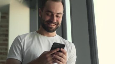 Evde cep telefonu kullanırken pencerenin yanında duran mutlu genç adam.
