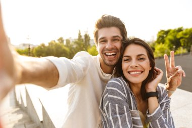 Parkta yürürken gülümseyen ve selfie çeken beyaz bir çiftin resmi.