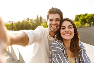 Parkta yürürken gülümseyen ve selfie çeken beyaz bir çiftin resmi.